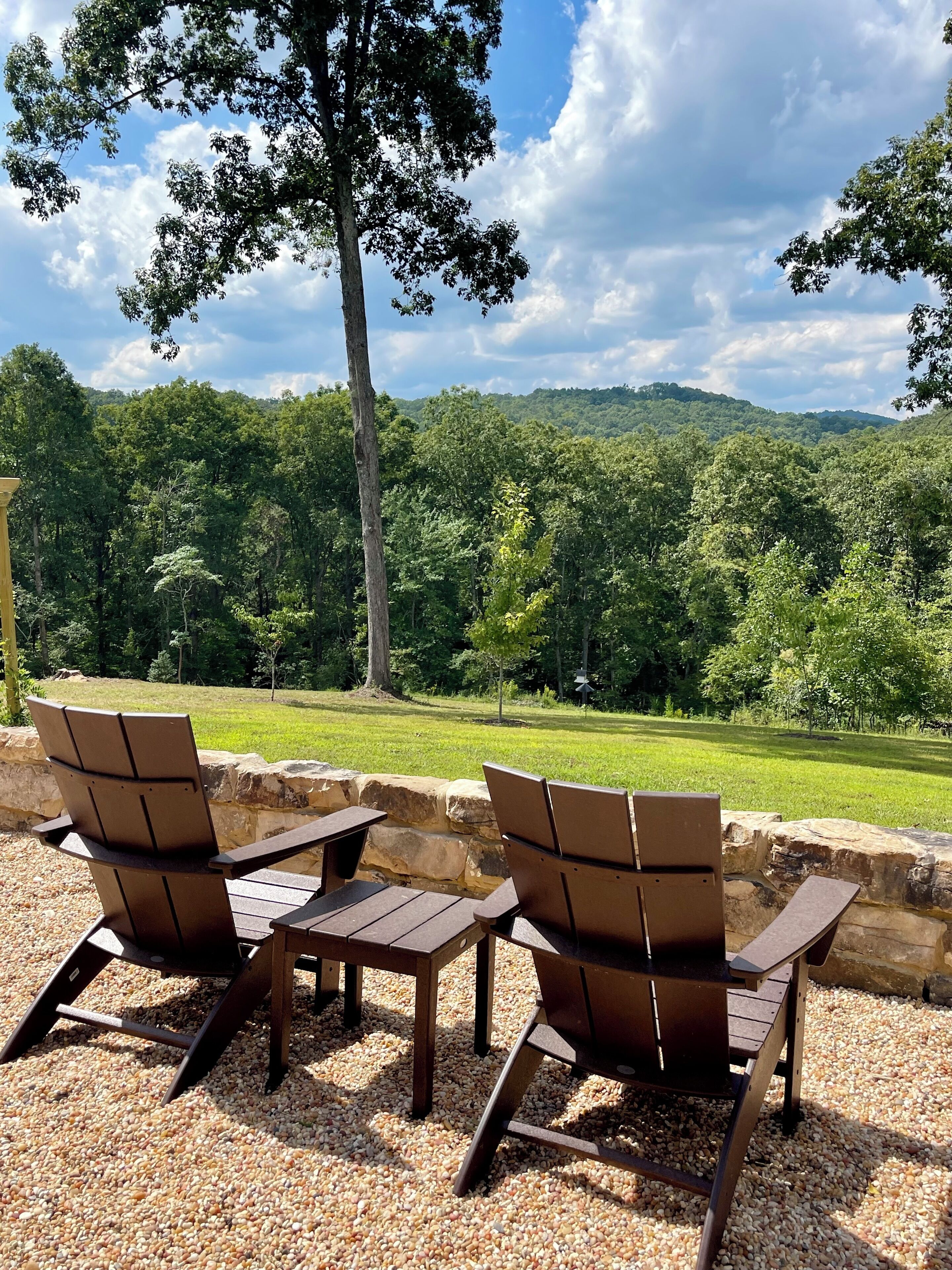 An outdoor seating area with Adirondack chairs and a table, surrounded by a stone wall and gravel, overlooking a lush green landscape and a forested hill.