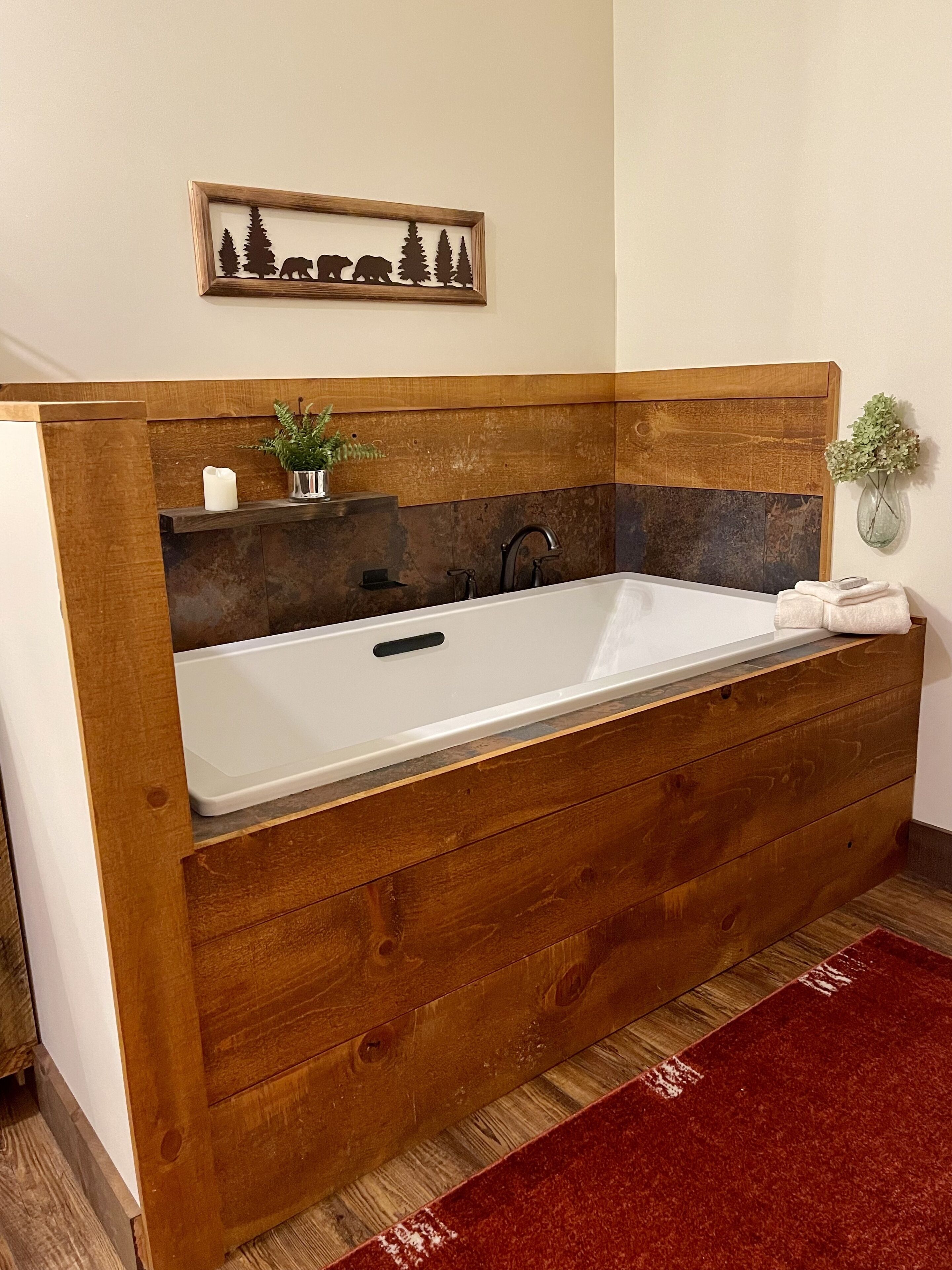 Ein Badezimmer mit einem Holzwaschtisch, einer weißen Badewanne, einem hölzernen Kopfteil, einer wandmontierten Pflanze und einem roten Teppich.