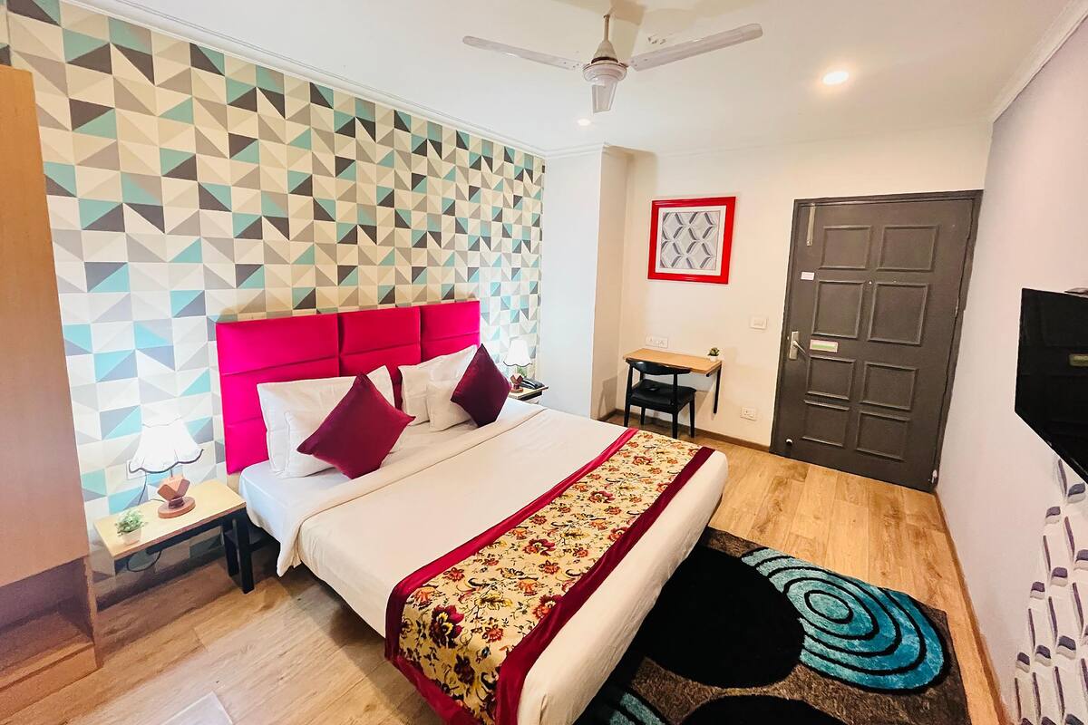 A hotel room with a geometric patterned wall, a red headboard, a bed with white and red bedding, a desk with a chair, a ceiling fan, and a door with a security lock.