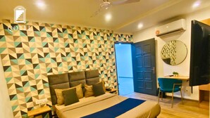 A hotel room with a geometric patterned wall, a bed with a blue bedspread, a desk with a chair, and a round mirror on the wall. - Qotel Comfort Chattarpur- Near Chattarpur Metro (New Delhi)