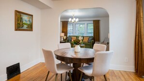 Ein Essbereich mit einem runden Tisch und sechs Stühlen, einem Bild an der Wand und Blick in ein Wohnzimmer mit Sofa und Fenster.