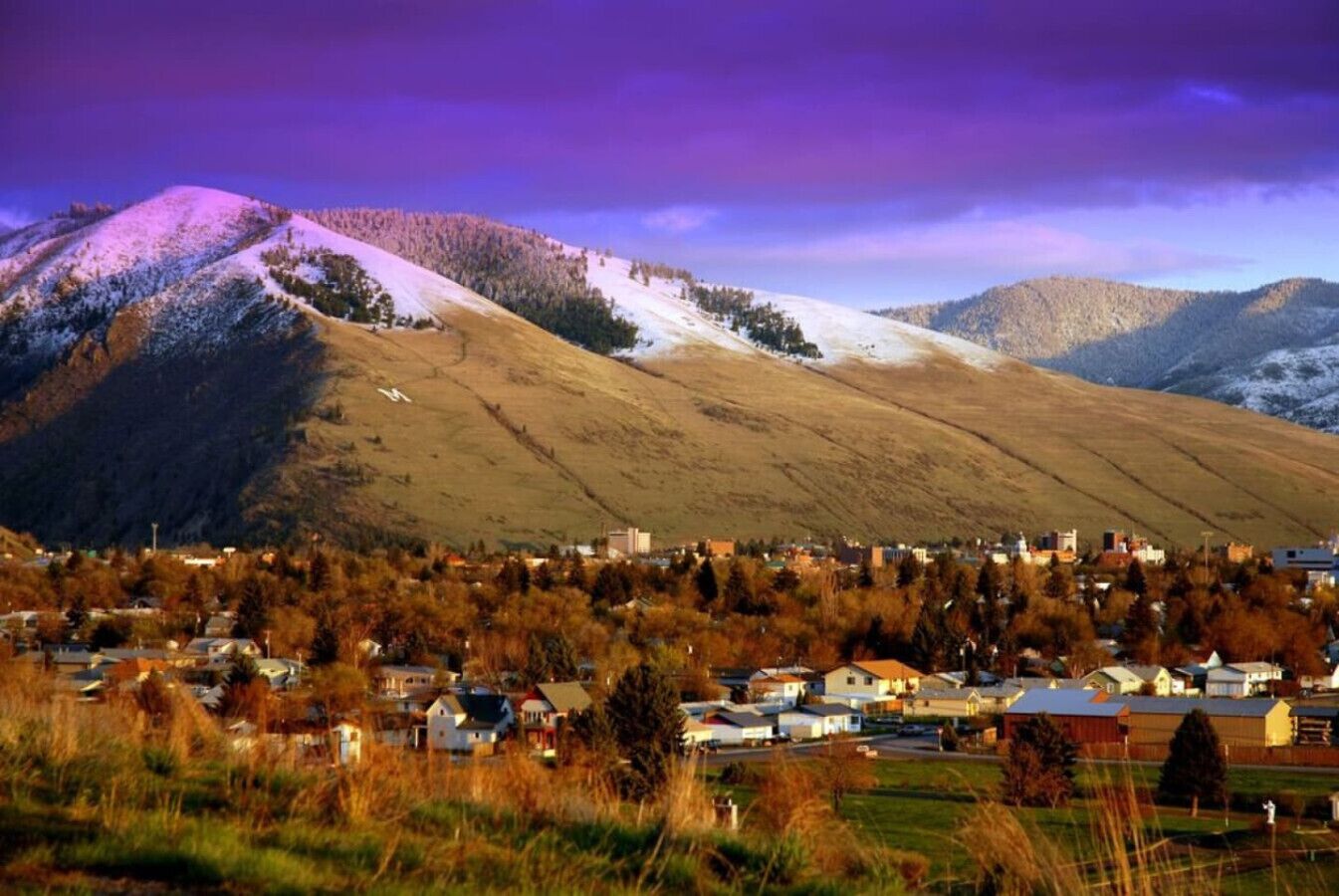A town nestled in a valley with a backdrop of snow-capped mountains and a purple sky.
