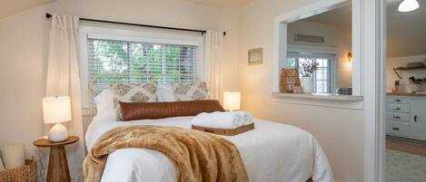 Une chambre à coucher avec un lit, des lampes de chevet, une fenêtre avec des stores, un miroir et une vue sur la salle de bain.