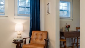 Un salon comprenant un fauteuil en cuir, une table d’appoint avec une lampe, une table à manger avec des chaises et une fenêtre ornée de rideaux bleus.