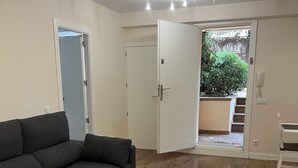 Een woonkamer met een donkergrijze bank, een witte salontafel en een grijze poef. Een witte deur leidt naar een balkon met uitzicht op groen.