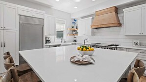 A modern kitchen with white cabinetry, a large island with a bowl of fruit, and a wooden range hood. - WaterColor PRIME Phase 1 Location! With NEW LSV & 4 Bikes! Walk to Seaside! (Santa Rosa Beach)