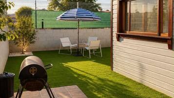 Un patio con césped artificial, una parrilla, una mesa con sillas y una cerca verde.