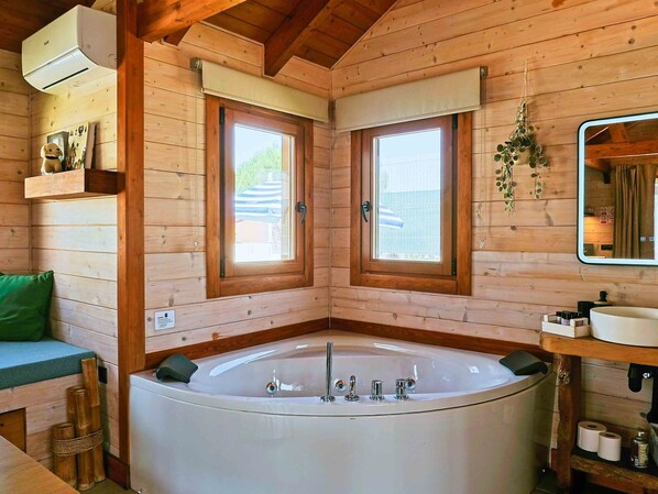 A wooden cabin interior with a large bathtub, two windows, a mirror, and a shelf with toiletries.