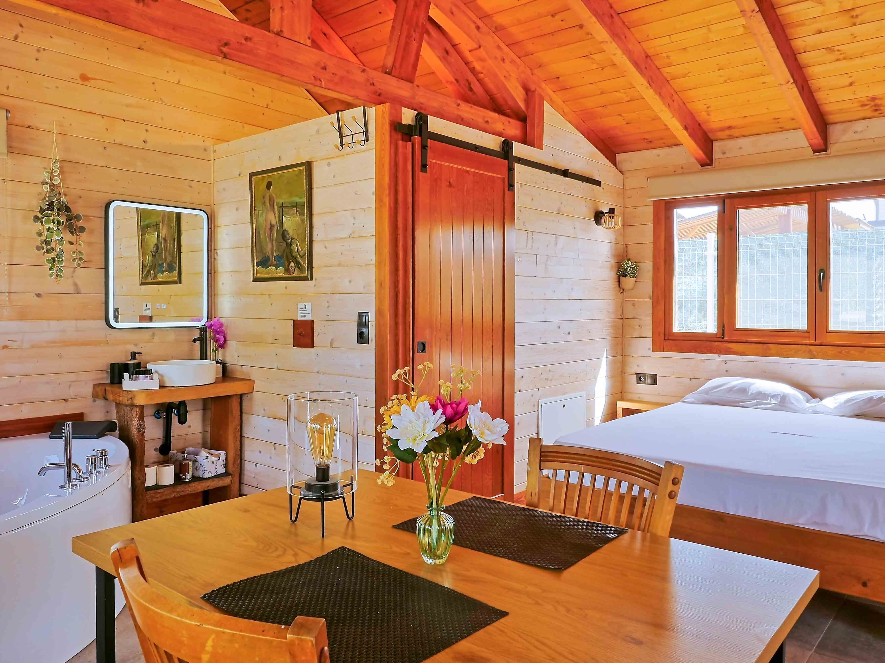 Una cabana de fusta amb un llit, una taula amb un ram de flors, un vàter i un mirall.