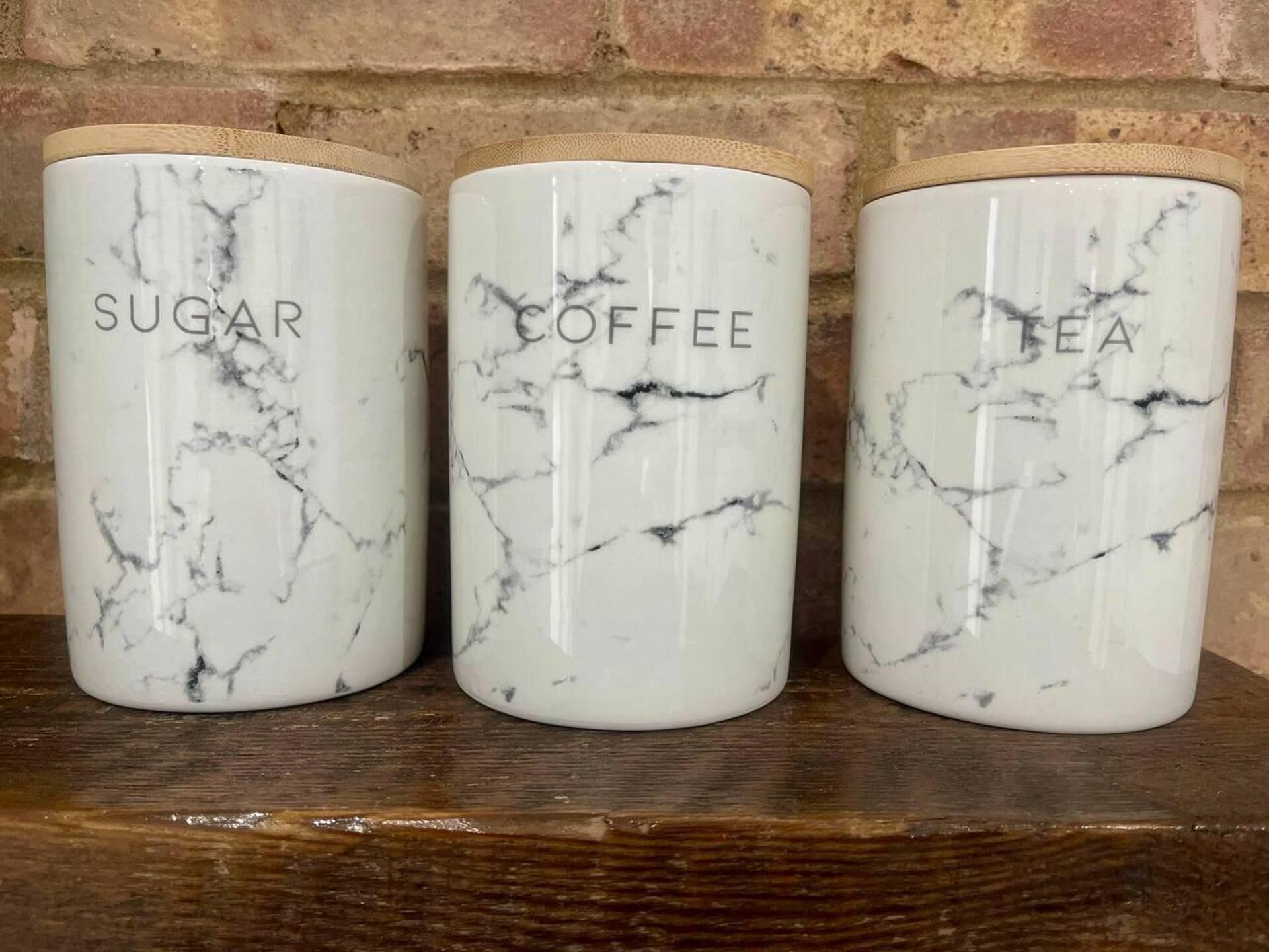 Three marble-patterned containers labeled 'SUGAR,' 'COFFEE,' and 'TEA' on a wooden shelf against a brick wall.