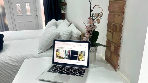 A laptop on a white table in a room with a bed, a brick wall, and a potted orchid. - Nice And Cosy Large Studio Near Luton Airport! (Luton)
