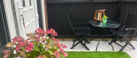 Une terrasse avec une table ronde et quatre chaises, une petite plante en pot et un gazon artificiel vert.