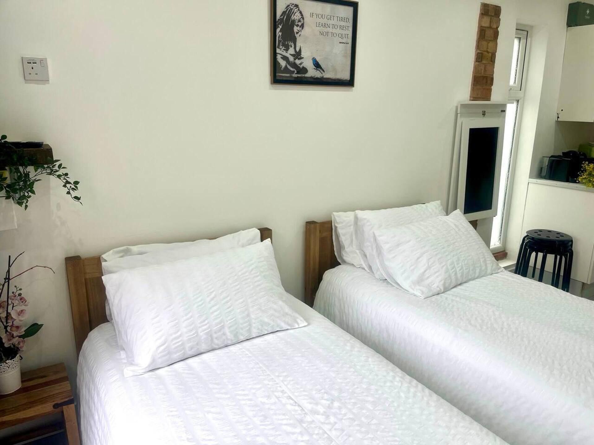 A bedroom with two single beds, a small stool, a potted plant, and a wall-mounted light switch.