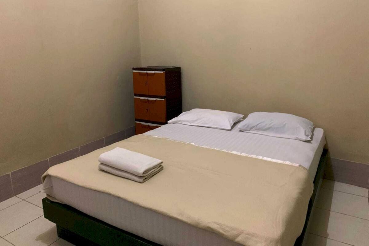 A small, neatly made bed with a beige bedspread and white pillows, a wooden dresser, and a tiled floor.