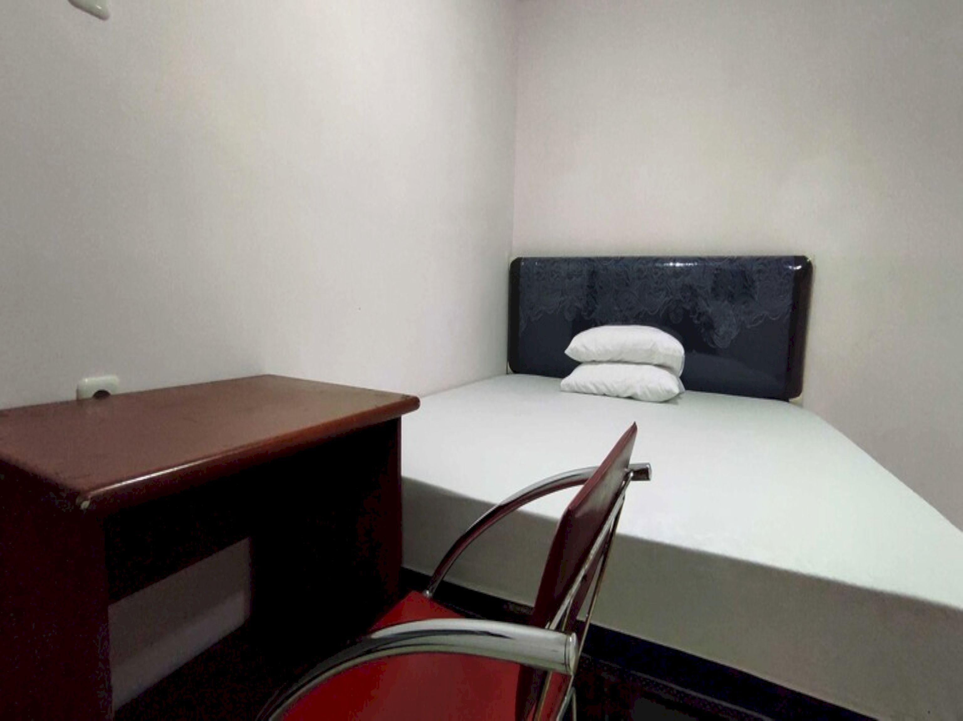 A small, single-bed room with a wooden desk and chair, a white mattress, and a dark headboard.