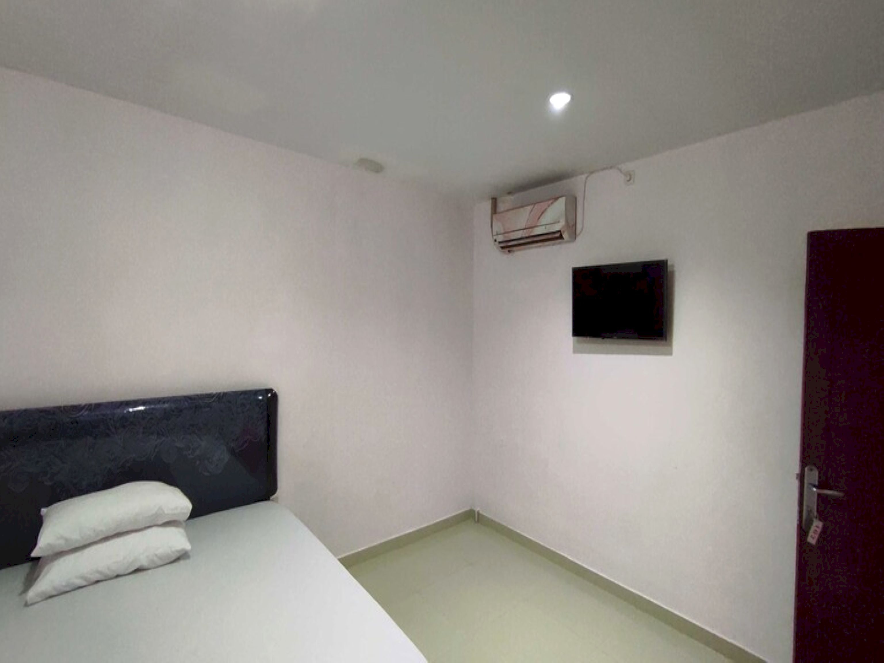 A small, sparsely furnished room with a bed, a wall-mounted air conditioner, a small TV, and a door.
