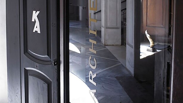 Une porte sur laquelle on peut lire les lettres « KA » et le mot « ARCHITETTURA », qui donne accès à un bâtiment à l’architecture classique et au sol en marbre.