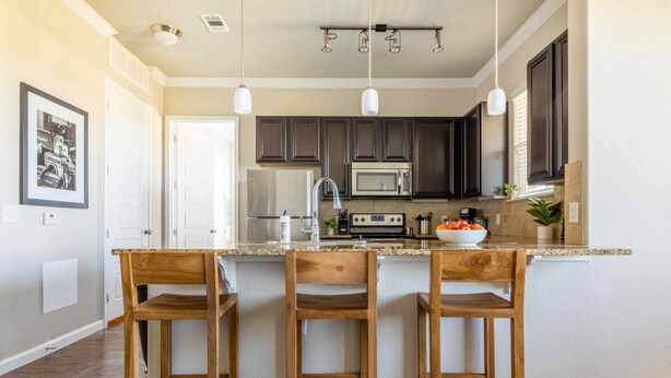 A kitchen with dark cabinets, a granite countertop, and three wooden bar stools. - Century Stone Hill North (Pflugerville)