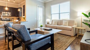 A modern living room with a sofa, coffee table, and chairs. There is a kitchen area with dark cabinets and a microwave. Large windows provide natural light. - Century Stone Hill North (Pflugerville)