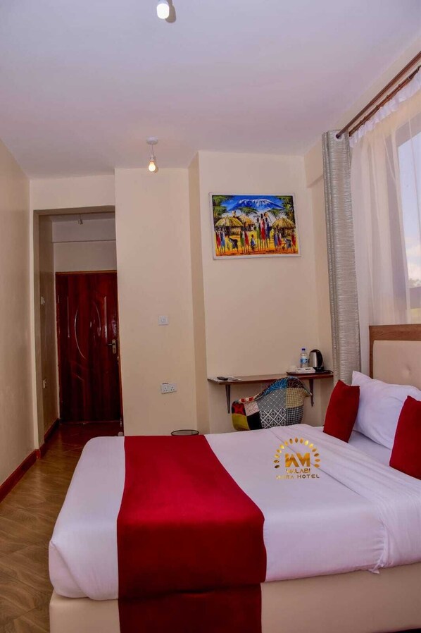 A hotel room with a bed, a painting on the wall, a desk with a chair, and a window with curtains.