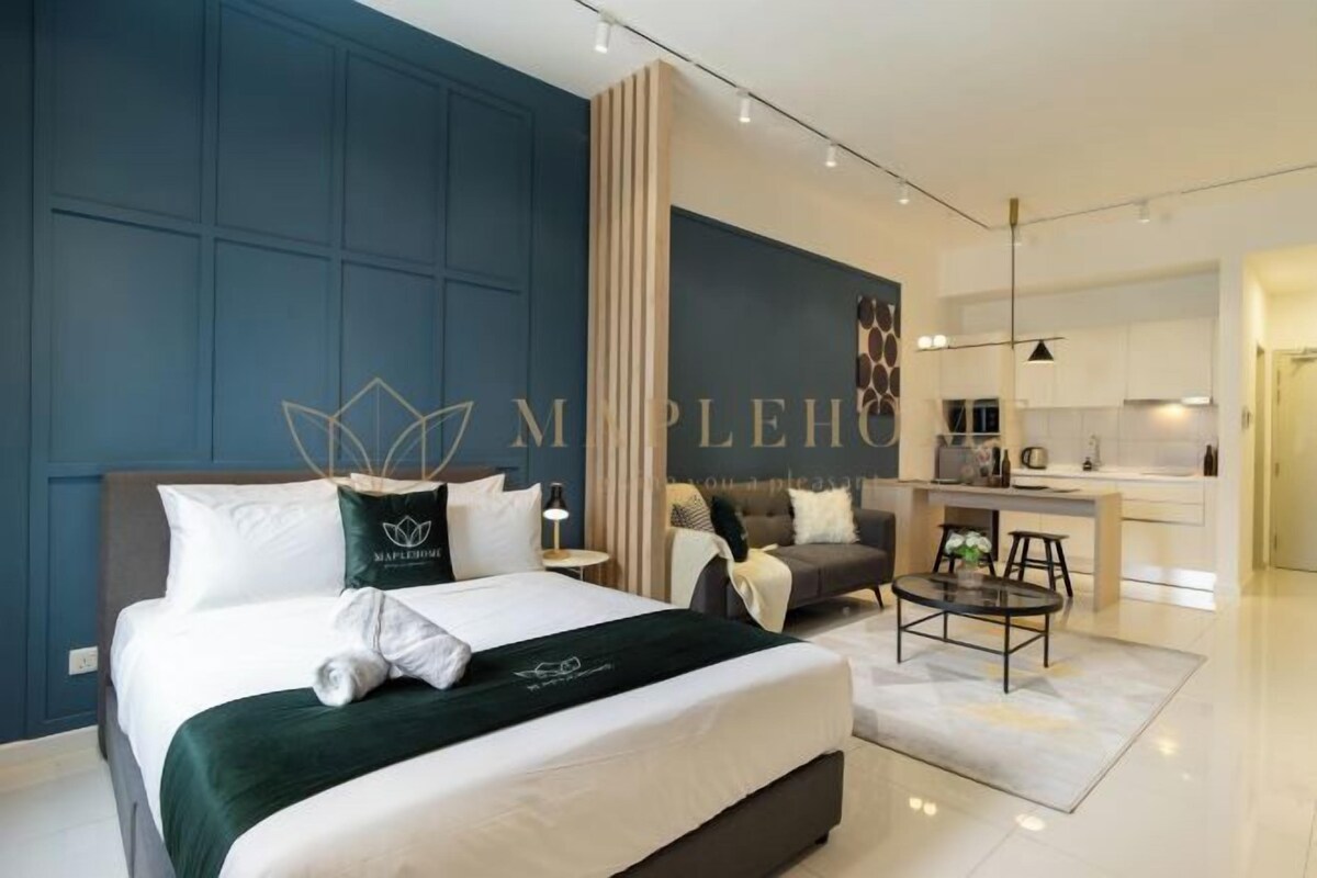 A modern hotel room with a large bed, a sitting area with a table, and a kitchenette in the background.