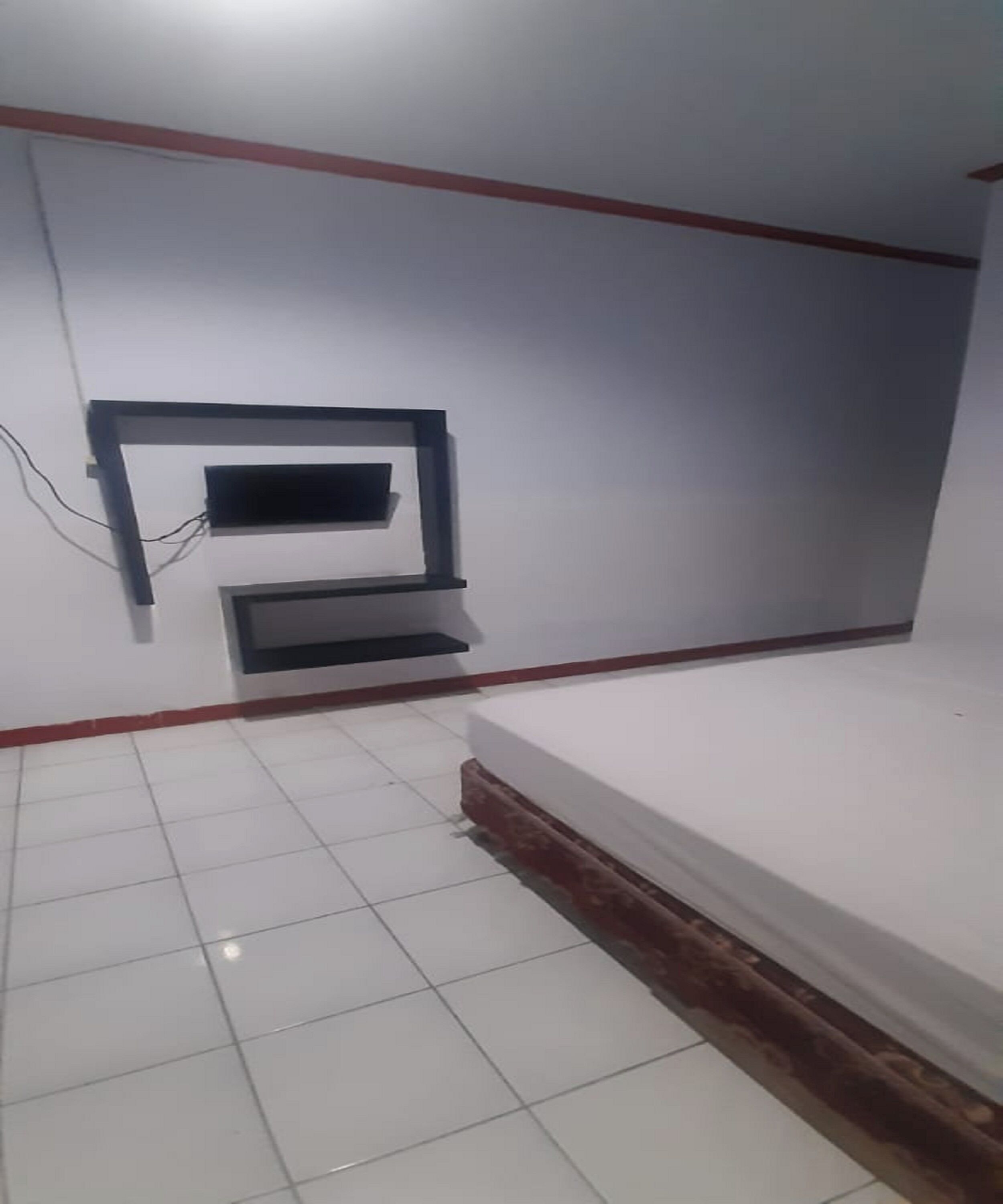 A room with a tiled floor, a bed, a wall-mounted TV, and a small shelf.