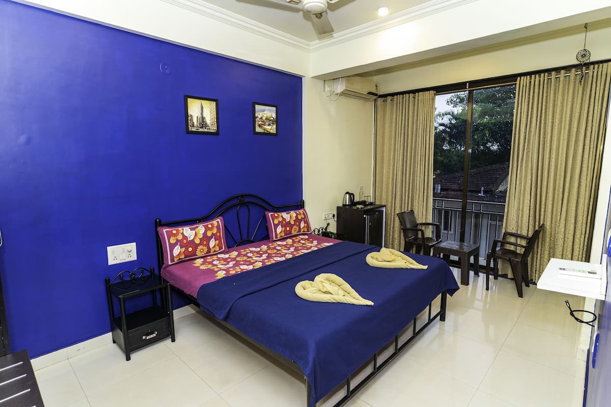 A hotel room with a blue accent wall, a bed with a blue bedspread, two pillows, a bedside table, a chair, a desk, and a window with curtains.