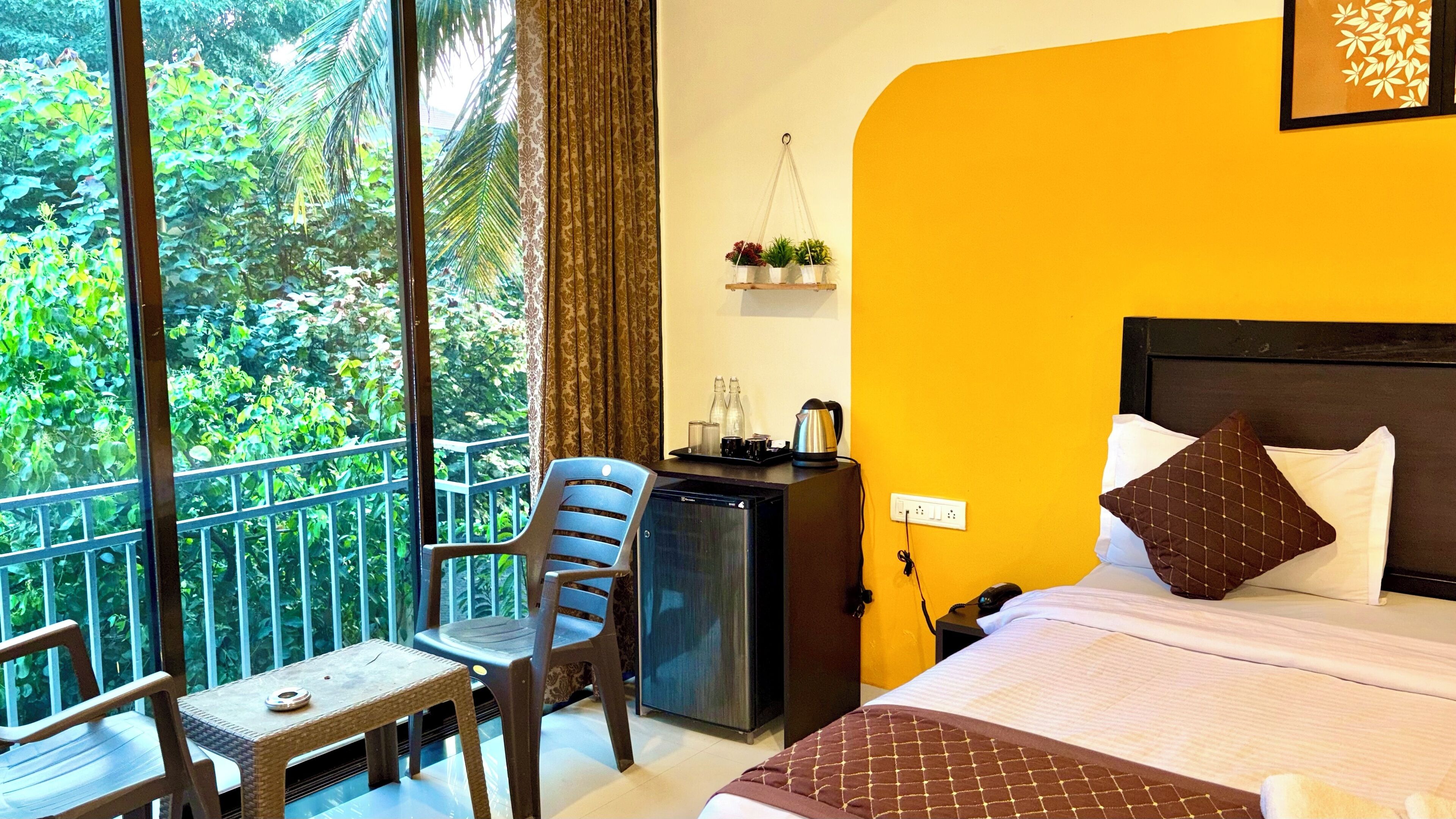 A hotel room with a bed, a desk with a computer, a chair, and a view of greenery through large windows.