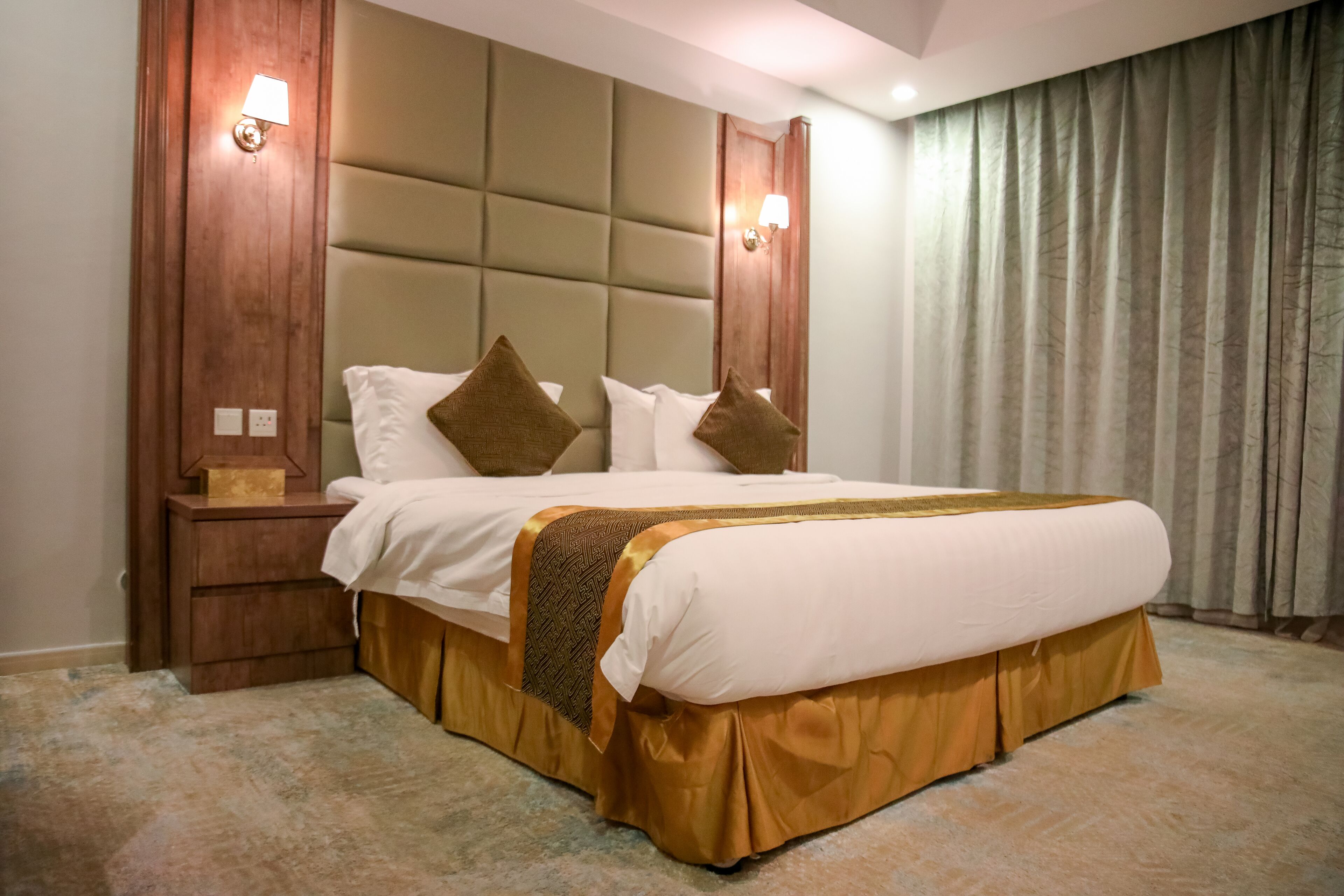 Habitación de hotel con una cama grande, cabecero de madera, dos lámparas de noche y piso cubierto de alfombra.