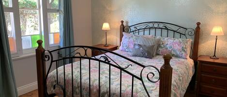Un dormitorio con estructura de cama metálica, ropa de cama floral, dos mesitas de noche con lámparas, una ventana con cortinas y un papel tapiz estampado.