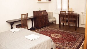 A room with a bed, a desk, two chairs, a dresser, a shelf with books, and a window with curtains.