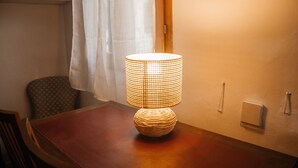 A table with a woven lamp, a chair, a bookshelf, and a wall-mounted light switch.