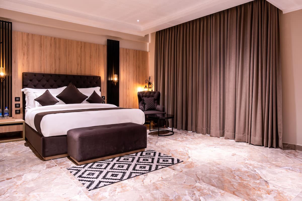 A modern bedroom with a large bed, a dark upholstered bench, a side table, a chair, and a patterned rug.