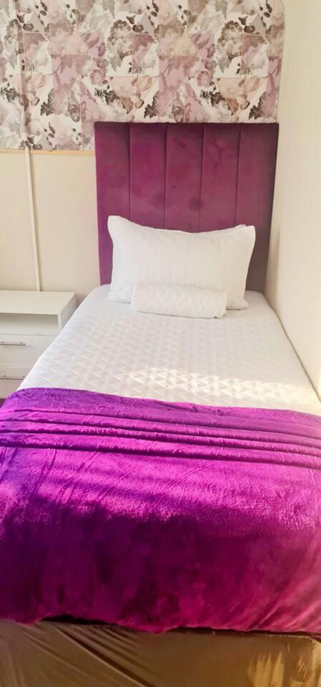 A bed with a purple headboard, white bedding, and a floral-patterned curtain.