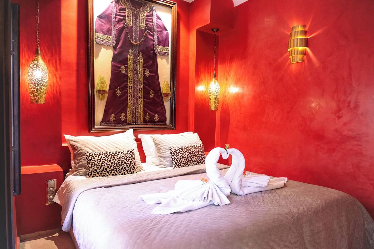 A hotel room with a red wall, a bed with white bedding, two pillows, a framed picture of a traditional outfit, and two wall-mounted lights.