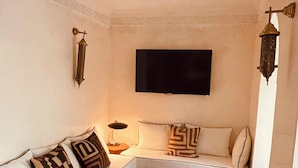 A living room with a white sofa, patterned cushions, a flat-screen TV, a small wooden table, and a woven rug. - Riad Oasissime Marrakech (Marrakech)