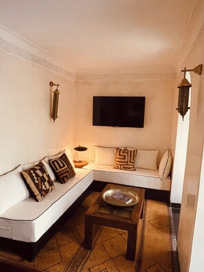 A living room with a white sofa, patterned cushions, a flat-screen TV, a small wooden table, and a woven rug. - Riad Oasissime Marrakech (Marrakech)