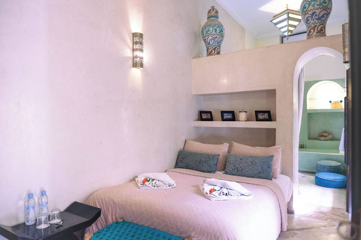 A bedroom with a bed, pillows, a nightstand, and a shelf with decorative items.