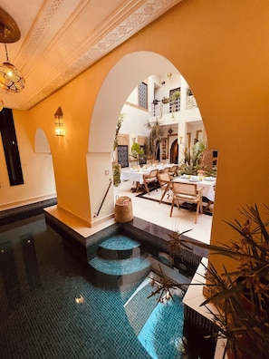 An indoor swimming pool with a tiled floor, a seating area with tables and chairs, and a decorative archway. - Riad Oasissime Marrakech (Marrakech)
