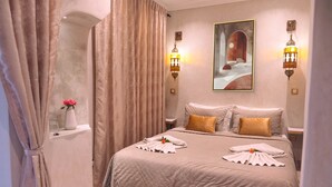 A hotel room with a large bed, beige curtains, a vase with a pink flower, and two wall-mounted lamps. - Riad Oasissime Marrakech (Marrakech)