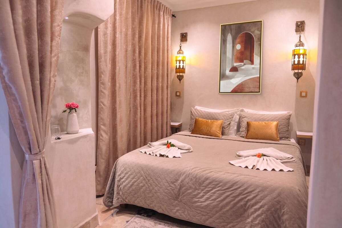 A hotel room with a large bed, beige curtains, a vase with a pink flower, and two wall-mounted lamps.