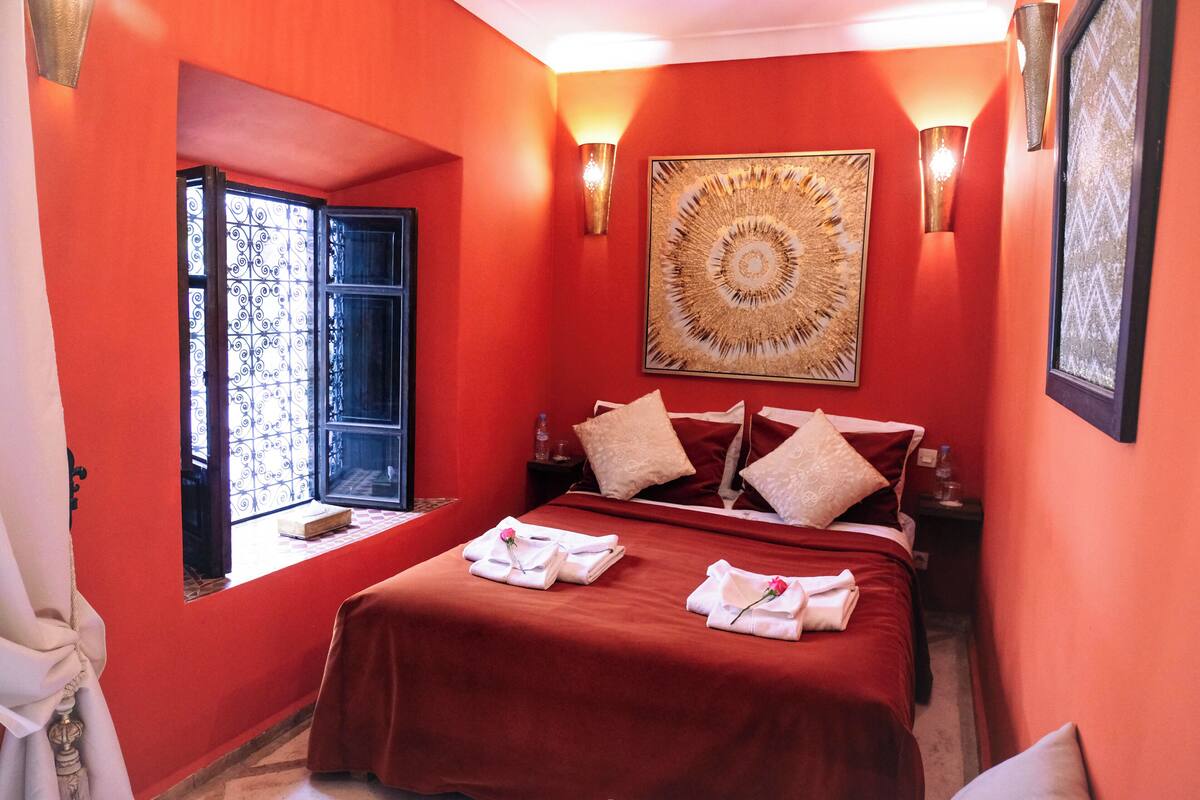 A hotel room with a red wall, a large bed with red bedding, two wall-mounted lamps, and a large abstract painting.