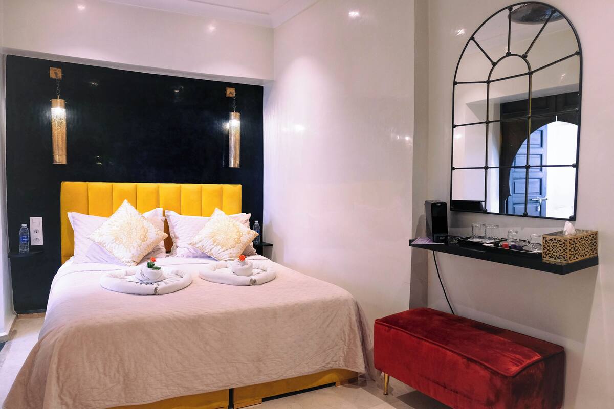 A modern hotel room with a large bed, a red bench, a black-framed mirror, and a small shelf with toiletries.