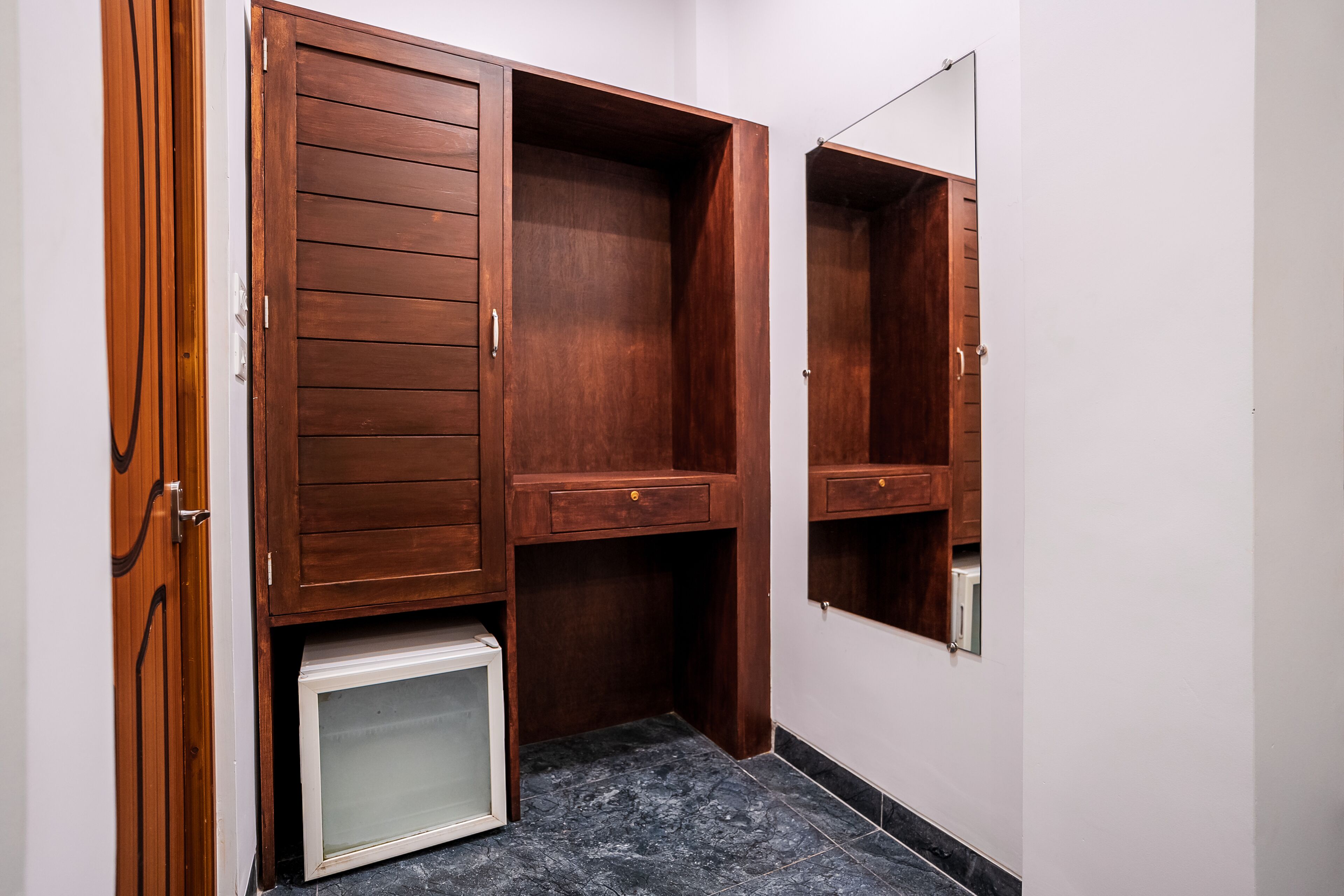 A compact room with a wooden wardrobe, a small white cabinet, and a mirror.