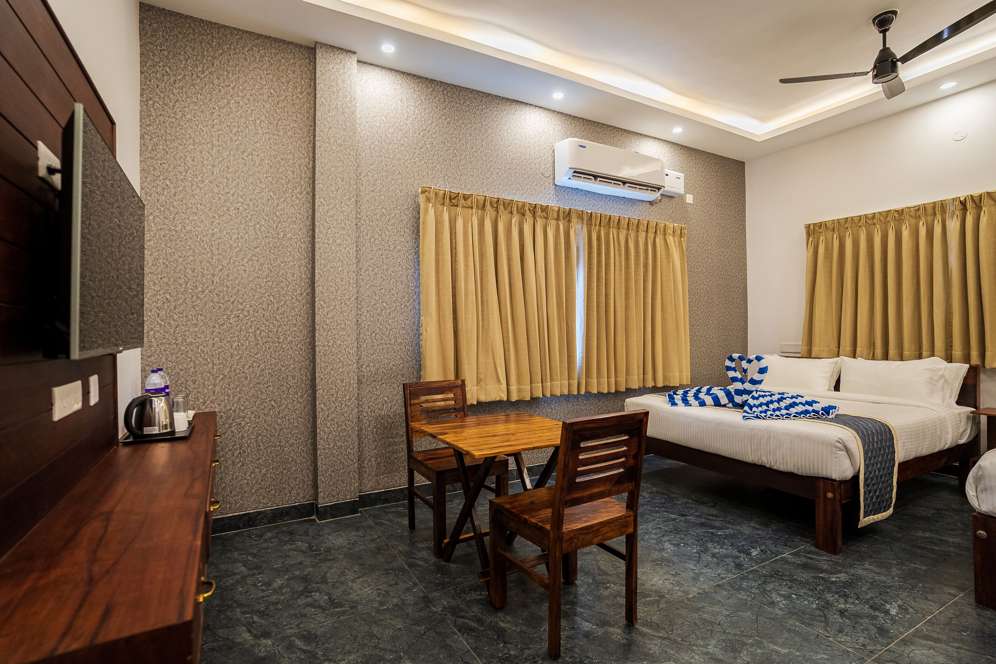 Quarto de hotel com cama, mesa de madeira com cadeiras, ar condicionado e ventoinha no teto.
