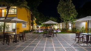 Un patio al aire libre con mesas y sillas, rodeado por un edificio amarillo y árboles.