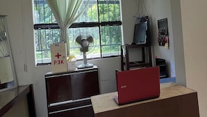 A room with a desk, a red laptop, a fan, a window with curtains, and a wall-mounted TV. - OYO 93178 Al Bait Homestay. (Lhokseumawe)