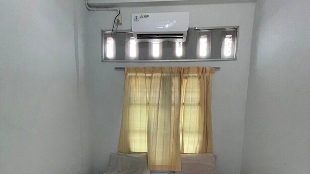 A small, single-bed room with a white bed, beige curtains, and a ceiling-mounted air conditioner. - OYO 93178 Al Bait Homestay. (Lhokseumawe)