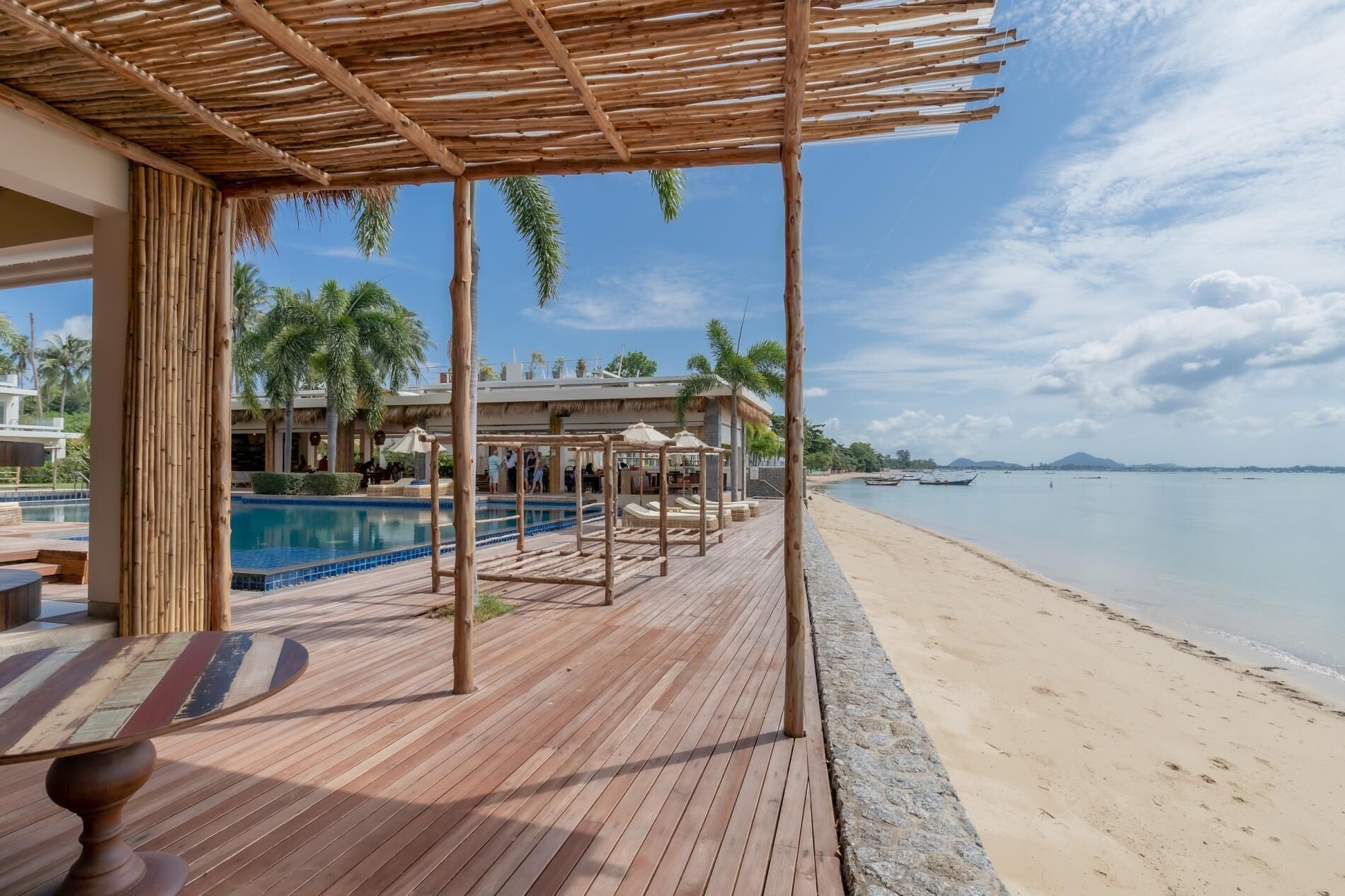 A wooden deck with a pool, beach, and boats in the background.