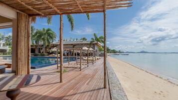 A wooden deck with a pool, beach, and boats in the background.